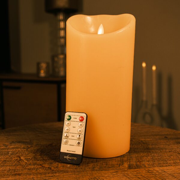 De Oplaadbare LED kaars D12,5 X H25 CM met flikkerend licht staan op een houten tafel naast een afstandsbediening, terwijl zacht gloeiende kaarsen en decor zorgen voor een warme sfeerverlichting op de achtergrond.