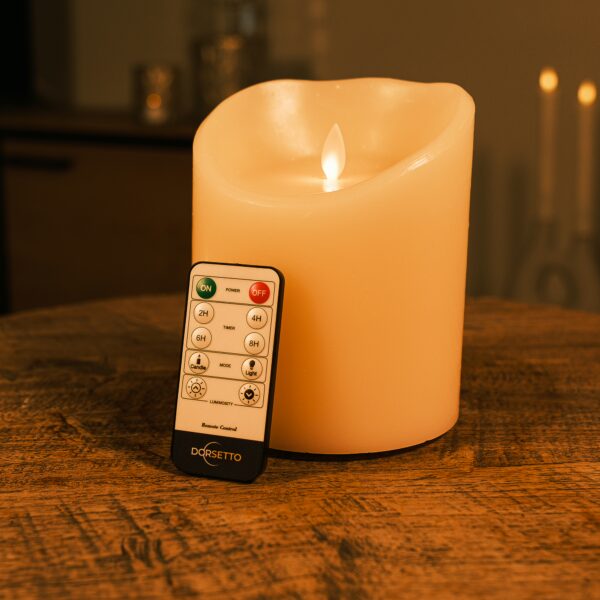 Een LED wax kaars D12,5 X H15 CM oplaadbaar warm wit met afstandsbediening gloeit op een houten tafel; de Dorsetio afstandsbediening ligt ervoor en zacht brandende vage kaarsen zijn zichtbaar op de achtergrond.