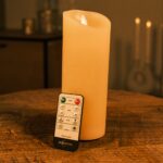 De LED wax kaars oplaadbaar DORSETTO D 7,5 X 20 CM staat op een houten tafel met de afstandsbediening dichtbij; zachte verlichting en vage kaarsen op de achtergrond dragen bij aan de gezellige sfeer.