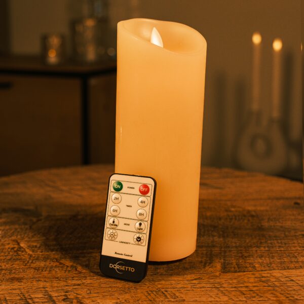 De LED wax kaars oplaadbaar DORSETTO D 7,5 X 20 CM staat op een houten tafel met de afstandsbediening dichtbij; zachte verlichting en vage kaarsen op de achtergrond dragen bij aan de gezellige sfeer.
