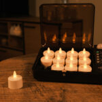 Twaalf LED-Waxine/theelichtjes waterdicht oplaadbaar brandtijd tot 400 uur warm wit (12 stuks) met afstandsbediening zitten in een zwarte oplaadkoffer op een houten tafel; één kaars staat buiten, met een zacht verlichte kamer op de achtergrond.