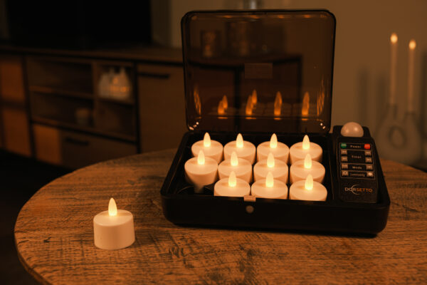 Twaalf LED-Waxine/theelichtjes waterdicht oplaadbaar brandtijd tot 400 uur warm wit (12 stuks) met afstandsbediening zitten in een zwarte oplaadkoffer op een houten tafel; één kaars staat buiten, met een zacht verlichte kamer op de achtergrond.