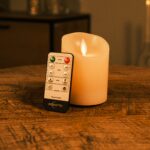 Een Dorsetto kaars met afstandsbediening staat rechtop voor een Oplaadbare LED wax kaars met bewegende vlam D 7,5 X 10 CM - warm wit op een houten tafel, met gezellige verlichting en vaag meubilair op de achtergrond.