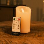 Een Oplaadbare LED wax kaars met bewegende vlam D 7,5 X 12,5 CM - warm wit staat op een houten oppervlak naast een DorsetTO afstandsbediening met timer en helderheidsknoppen en werpt een gezellig warm licht.
