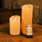 Twee Oplaadbare LED wax kaarsen set van 2 DORSETTO van verschillende hoogtes staan gloeiend op een houten tafel. Een DORSETTO-afstandsbediening ligt ervoor, met een zacht verlichte achtergrond voor extra sfeer.