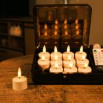 Een set van 12 oplaadbare LED waxinelichtjes 3D (tot 400 uur brandtijd) met timer en afstandsbediening, een weergegeven op een houten tafel. De warme verlichting creëert een gezellige sfeer, terwijl de afstandsbediening in de kist rust.
