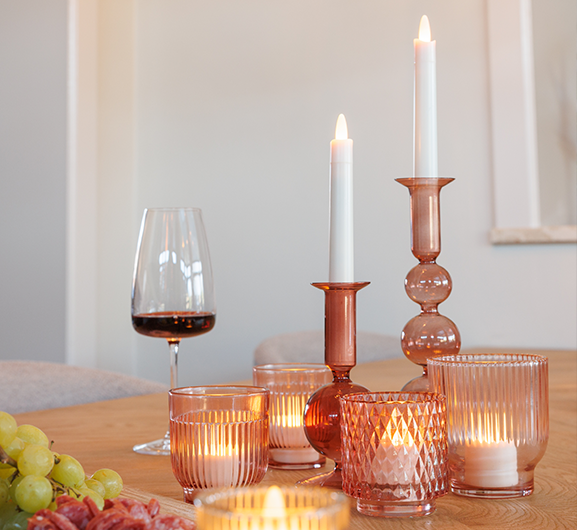Een gedekte tafel met brandende kaarsen in roze glazen houders, een glas rode wijn en een tros groene druiven. Het zachte kaarslicht creëert een warme, uitnodigende sfeer.
