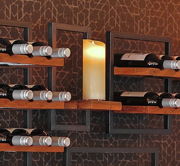 Wijnflessen staan horizontaal op houten rekken die zijn bevestigd aan een muur met een geometrisch patroon. Een vlamloze LED-kaars staat op een van de rekken en creëert een gezellige sfeer.
