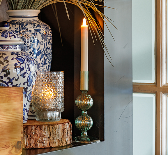 Een brandende conische kaars in een groene glazen houder staat naast een facetgeslepen glazen votief met een brandende kaars, beide geplaatst op een houten schijf naast een grote blauwwitte bloemenvaas met hoog gras.