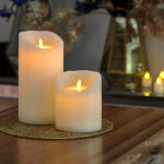 De LED wax kaarsen set 2 maten warm wit oplaadbaar DORSETTO met gebogen toppen staat op een geweven gouden placemat op een houten tafel, omringd door meer kaarsen en vazen met decoratief gebladerte voor een warme, gezellige sfeer.