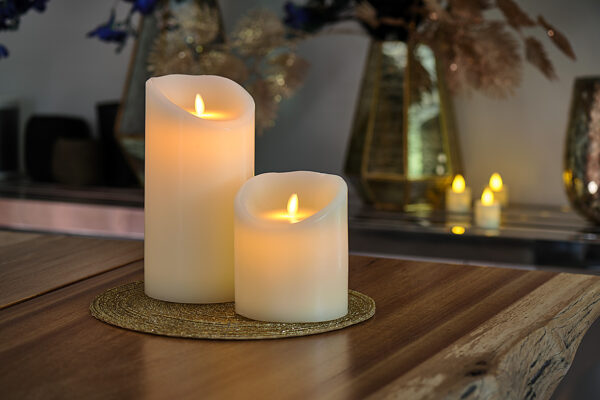 De LED wax kaarsen set 2 maten warm wit oplaadbaar DORSETTO met gebogen toppen staat op een geweven gouden placemat op een houten tafel, omringd door meer kaarsen en vazen met decoratief gebladerte voor een warme, gezellige sfeer.