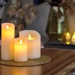 Een Oplaadbare LED wax kaars met bewegende vlam D 7,5 X 12,5 CM - warm wit zit op een geweven mat op een houten tafel, met zacht gloeiende decoratieve vazen en kaarsen vervaagd op de achtergrond.