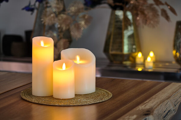 Een Oplaadbare LED wax kaars met bewegende vlam D 7,5 X 12,5 CM - warm wit zit op een geweven mat op een houten tafel, met zacht gloeiende decoratieve vazen en kaarsen vervaagd op de achtergrond.