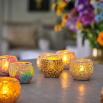 Zeven Waxinelichthouder 9 CM Dorsetto mozaïek multicolor glazen waxinelichthouders gloeien in levendige kleuren op een tafel, terwijl vage kleurrijke bloemen en een bank op de achtergrond bijdragen aan de gezellige, decoratieve sfeer.