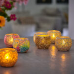 Zes kandelaars van mozaïekglas, waaronder de Waxinelichthouder 8,5 CM Dorsetto mozaïek rood, met brandende waxinelichtjes staan op een tafel; sommige zijn goudkleurig en andere kleurrijk. Een wazig boeket bloemen en meubels verschijnen op de achtergrond.