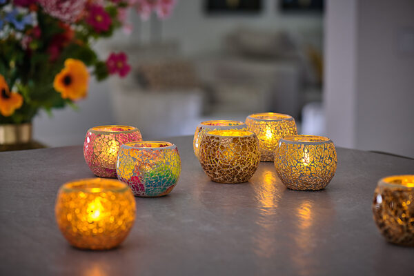 Twaalf waterdichte, oplaadbare waxinelichtjes met afstandsbediening (brandduur 175 uur, warm wit) gloeien in mozaïekglazen houders met gouden en kleurrijke patronen. Op de onscherpe achtergrond staan bloemen en een gezellige woonkamer.