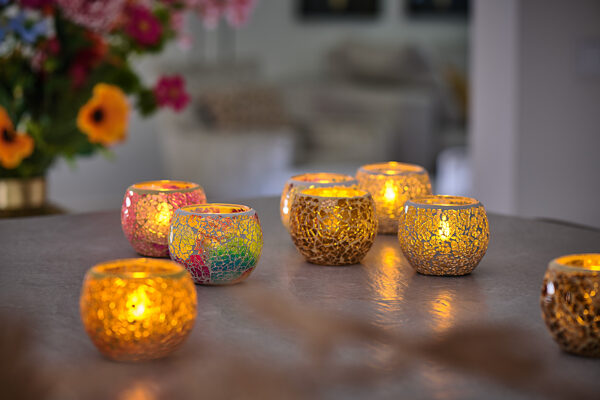 De Waxinelichthouder 8,5 CM Dorsetto mozaïek rood, met een brandende kaars erin, staat uitgestald tussen andere kleurrijke mozaïek glazen houders op een tafel. Op de zacht vervaagde achtergrond staan bloemen en een warme, gezellige omgeving binnenshuis.