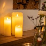 De LED wax kaarsen set 3 kaarsen warm wit oplaadbaar DORSETTO staat op een houten oppervlak bij een witte muur, met vage glazen flessen en bloemenvazen op de voorgrond, die een gezellige en uitnodigende gloed aan het tafereel toevoegen.