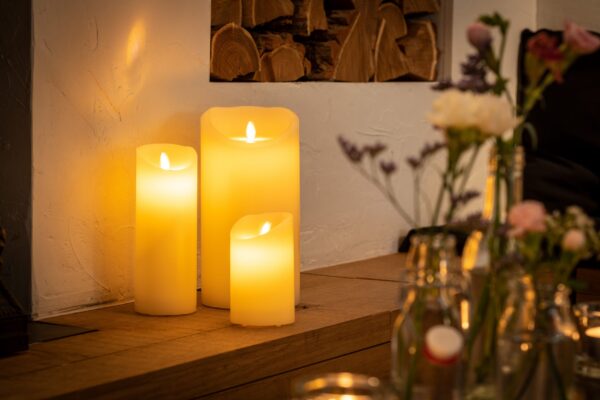 De LED wax kaarsen set 3 kaarsen warm wit oplaadbaar DORSETTO staat op een houten oppervlak bij een witte muur, met vage glazen flessen en bloemenvazen op de voorgrond, die een gezellige en uitnodigende gloed aan het tafereel toevoegen.