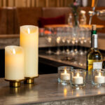 Twee Oplaadbare LED wax kaarsen met bewegende vlam (7,5 x 12,5 cm, warm wit) en drie kleine votiefkaarsen gloeien op de bar. Wijnflessen, glazen en bloemenvazen voegen een gezellige elegantie toe aan het tafereel.