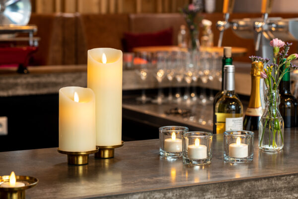 Twee Oplaadbare LED wax kaarsen met bewegende vlam (7,5 x 12,5 cm, warm wit) en drie kleine votiefkaarsen gloeien op de bar. Wijnflessen, glazen en bloemenvazen voegen een gezellige elegantie toe aan het tafereel.