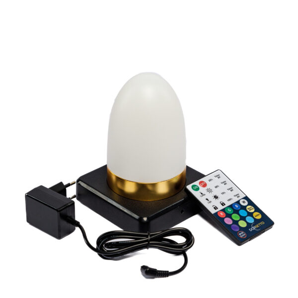 De Tafellamp DORSETTO is een oplaadbare, waterdichte tafellamp (105 uur), ovaal met witte kap en goudkleurige basis. Hij staat op een zwart platform, met afstandsbediening met gekleurde knoppen en zwarte adapter ernaast.