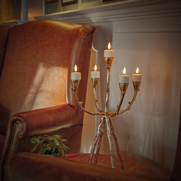 Een gouden kandelaar met zes LED waxinelichtjes 3D (brandtijd 175 uur, oplaadbaar, met afstandsbediening & timerfunctie) staat op een houten tafel tussen twee bruinleren fauteuils; een kleine potplant staat ernaast.