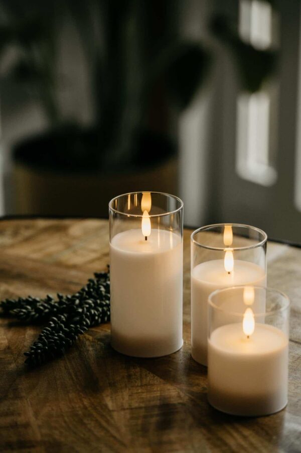 Drie DORSETTO kaarsen in glas (oplaadbaar, 105 uur brandtijd, 3D vlam, met afstandsbediening) staan aan in glazen houders op hout, met groen ernaast. De onscherpe achtergrond zorgt voor een warme en gezellige sfeer.