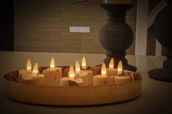 Een rond dienblad toont zes 3D oplaadbare LED-was/theelichtjes (175 uur, met afstandsbediening, timerfunctie), hun warme gele licht creëert een gezellige sfeer. Twee donkere kaarsenhouders staan op de achtergrond op een glad oppervlak.