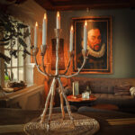 Een zilveren kandelaar met zes LED dinerkaarsen oplaadbaar 97mm 130 uur warm wit (set van 6, met afstandsbediening) staat op een geweven mat in een gezellige kamer en werpt warm licht bij een bank en een groot ingelijst portret.