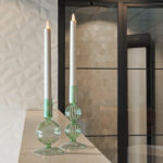 Twee hoge witte LED dinerkaarsen 270mm oplaadbaar uit een 6-delige set met afstandsbediening, die 130 uur lang warm wit opgloeien, staan in elegante groene houders op een licht aanrecht, tegen geometrische witte tegels en een glazen deur.