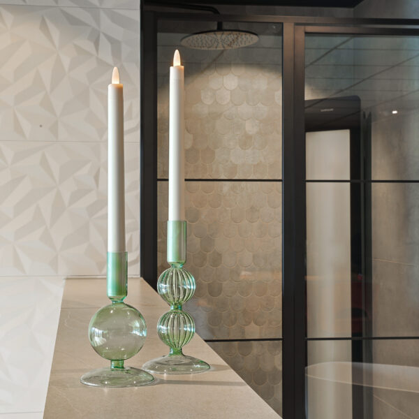 Twee brandende Oplaadbare LED dinerkaarsen 270mm 130 uur warm wit rusten in groene glazen houders op een beige oppervlak, met geometrische en geschulpte wandtegels plus een douchewand van zwart omlijst glas op de achtergrond.