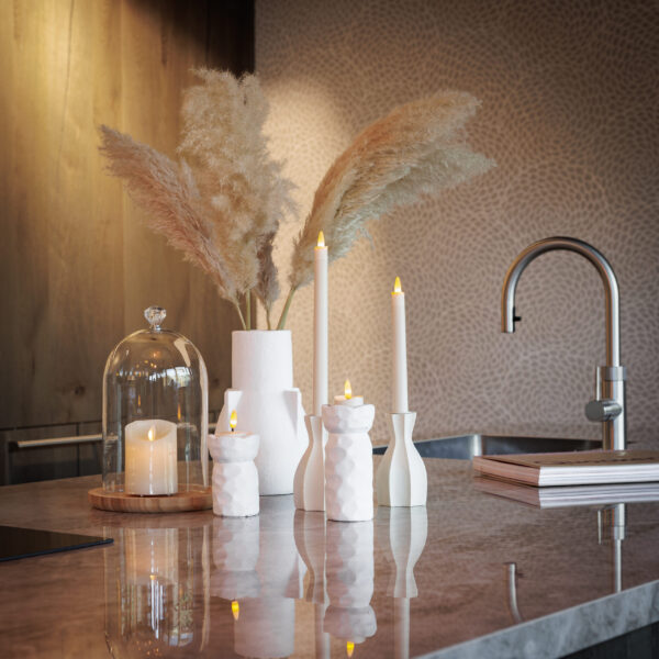 Een modern aanrecht toont witte keramische kandelaars met LED waxinelichtjes 3D (brandtijd 175 uur, oplaadbaar, afstandsbediening, timerfunctie, set van 6), een glazen cloche boven een kaars, pampagras in een vaas en zachte neutrale verlichting voor gezelligheid.