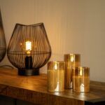 Drie DORSETTO oplaadbare led kaarsen (set van 3, brandtijd 105 uur, incl. afstandsbediening) in glazen houders staan op hout naast metalen lampen-perfect voor hotels en direct warme sfeer na opladen.