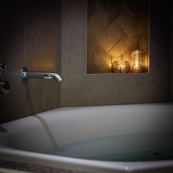 Een modern bad met water staat bij een bruin betegelde muur. Een ingebouwde plank erboven bevat Oplaadbare LED waxine/theelichtjes 175 uur waterproof warm wit (6 stuks) met afstandsbediening in glazen houders, waardoor een gezellige, ontspannen sfeer wordt gecreëerd.