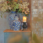 Blauw en wit gedessineerde keramische vazen met witte bloemen en groen staan op een houten oppervlak naast een Oplaadbare LED wax kaars met bewegende vlam D 7,5 X 10 CM - warm wit in een zwarte houder, tegen betegelde en betonnen muren.