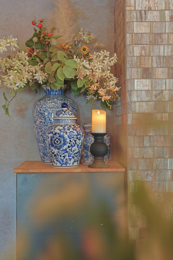 Blauw en wit gedessineerde keramische vazen met witte bloemen en groen staan op een houten oppervlak naast een Oplaadbare LED wax kaars met bewegende vlam D 7,5 X 10 CM - warm wit in een zwarte houder, tegen betegelde en betonnen muren.