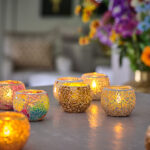 Een levendige Waxinelichthouder 8,5 CM Dorsetto mozaïek rood met brandende kaarsen staat op een tafel, omringd door andere kleurrijke kandelaars. Een bloemenboeket en een bank met patroon verschijnen zachtjes op de achtergrond en creëren een warme, uitnodigende sfeer.