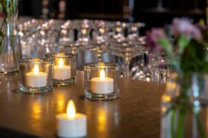 Twaalf Led waxinelichtjes waterdicht oplaadbaar brandtijd 175 uur warm wit met afstandsbediening staan in heldere houders op een aanrecht, met onscherp glaswerk en bloemen op de achtergrond, waardoor een warme, uitnodigende sfeer wordt gecreëerd.