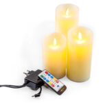 Drie Oplaadbare LED wax kaarsen van 3 stuks DORSETTO in verschillende hoogtes staan bij elkaar en stralen een warmgeel licht uit. Een afstandsbediening en stroomadapter zijn vooraan geplaatst.