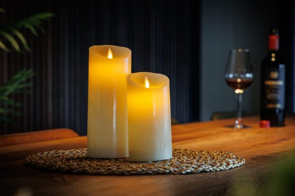 Twee gloeiende Oplaadbare LED wax kaarsen set van 2 DORSETTO zitten op een geweven placemat op een houten tafel, met een glas rode wijn en een fles op de zacht verlichte achtergrond, waardoor een uitnodigende en gezellige sfeer ontstaat.