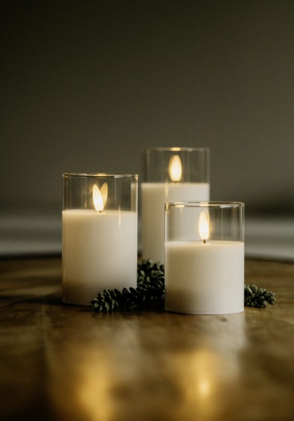 Drie DORSETTO led kaarsen uit de oplaadbare set (105 uur, 3D vlam, met afstandsbediening) branden in heldere glas houders op hout, omringd door groene sprietjes en creëren een warme, gezellige sfeer.