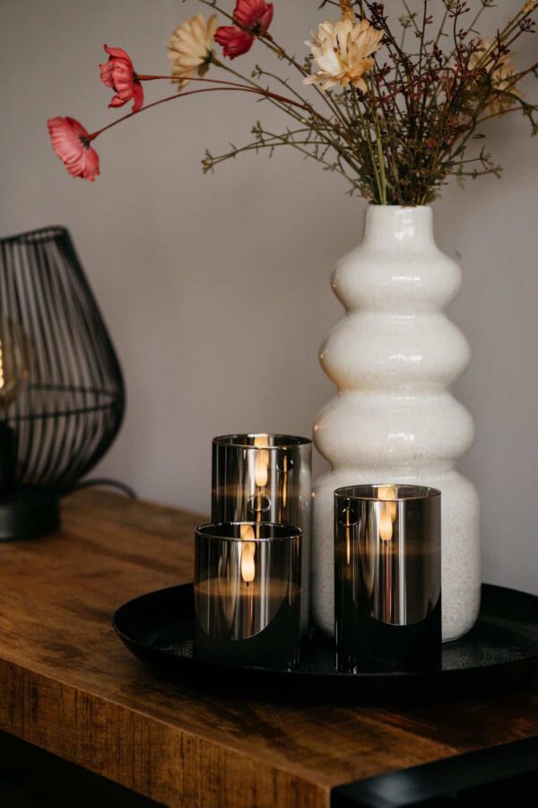 Een witte vaas met droogbloemen en de DORSETTO oplaadbare led kaarsen set 105 uur (3D vlam, afstandsbediening) staan op een ronde tray op hout. Op de achtergrond is deels een moderne zwarte draadlamp zichtbaar.