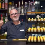 Een glimlachende barman met bril steekt zijn duim omhoog achter een met flessen beklede bar. Voor hem staat de Oplaadrek DORSETTO 35,5X21X51cm, een displaystandaard van drie niveaus met LED-kaarsen op batterijen.