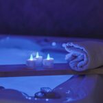 Drie RGB LED-Waxine/theelichtjes (waterdicht, 68 uur, oplaadbaar, 6 stuks met afstandsbediening) gloeien op een houten plank naast een opgerolde witte handdoek boven een zacht verlichte blauwe badkuip voor een ontspannen spasfeer.