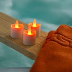 Twaalf oplaadbare, waterdichte RGB LED-Waxine/theelichtjes met afstandsbediening zitten op een houten richel naast een gevouwen oranje handdoek en creëren een uitnodigende spa- of zwembadsfeer.