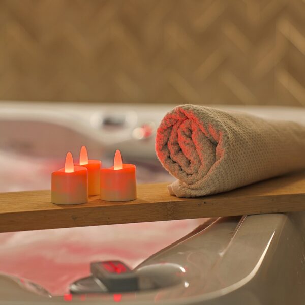 Een opgerolde beige handdoek en RGB LED-Waxine/theelichtjes waterdicht 68 uur oplaadbaar (6 stuks) met afstandsbediening gloeien op een houten plank boven een gevulde badkuip en creëren zo een ontspannen spa-achtige sfeer.