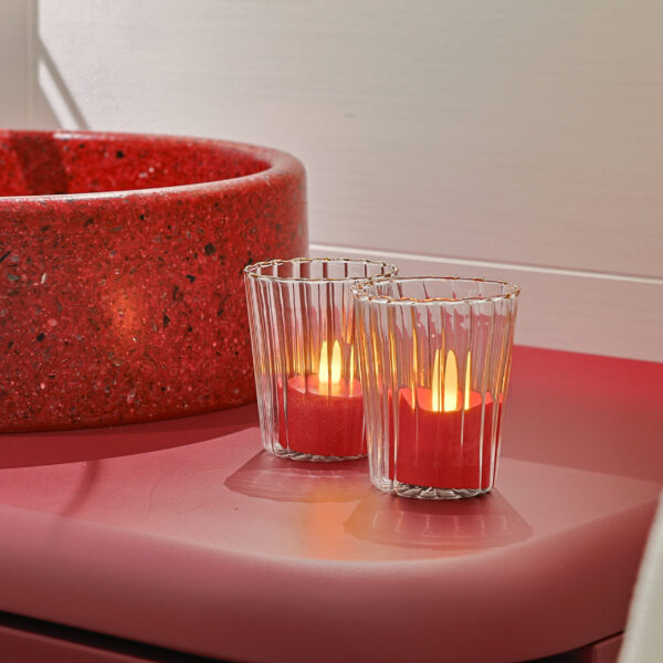 Twee Oplaadbare LED-kaarsjes rood - waterdicht - met Afstandsbediening (6 stuks, timerfunctie) gloeien warm op in geribbelde glazen houders op een roze oppervlak naast een ronde, roodgespikkelde schaal, waardoor een gezellige sfeer ontstaat.