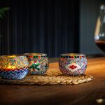 Drie kleurrijke Waxinelichthouder 9 CM Dorsetto regenboogkandelaars met gloeiende kaarsen staan op een geweven mat naast een glas rode wijn en creëren een warme, gezellige sfeer met zachte verlichting en een plant op de achtergrond.