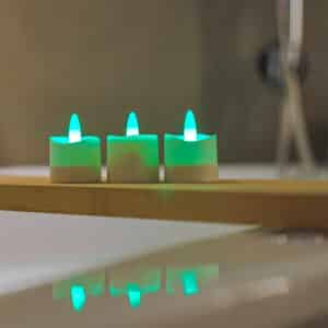 Drie kleine, witte RGB Led Waxinelichtjes oplaadbaar waterdicht (set van 12) met afstandsbediening gloeien groen op een houten dienblad, hun reflecties zichtbaar op het gladde oppervlak eronder tegen een zacht wazige achtergrond.